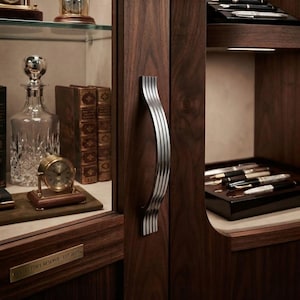 May include: A dark wood display cabinet featuring a vertical silver metal handle with a ribbed design. The cabinet interior holds a glass decanter, a small gold-toned desk clock, leather-bound books, and a tray of fountain pens. A brass plaque reads COLLECTOR'S RESERVE - EST 2021.