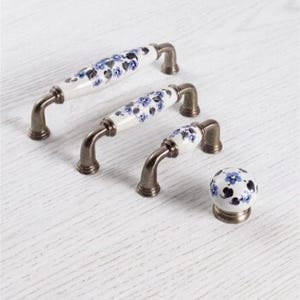 May include: A collection of four cabinet hardware pieces including three arched handles and one round knob. Each piece features a white ceramic center with a blue floral pattern and dark metallic bronze-toned metal ends and bases.
