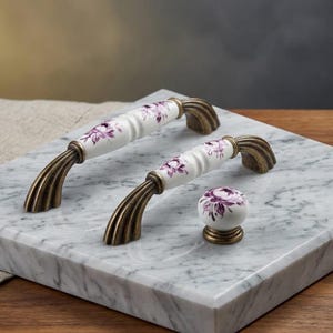 May include: Two arched cabinet pulls and one round cabinet knob made of white ceramic with a purple floral pattern. Each piece features an antique brass finish on the metal mounting hardware, which has a ribbed, fluted design at the base.