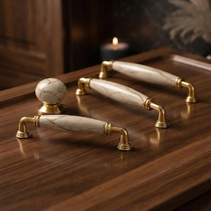 May include: A set of three cabinet hardware pieces including two elongated drawer pulls and one round knob. Each piece features a polished gold-tone metal base and a central beige stone-like insert with subtle brown veining throughout the surface.