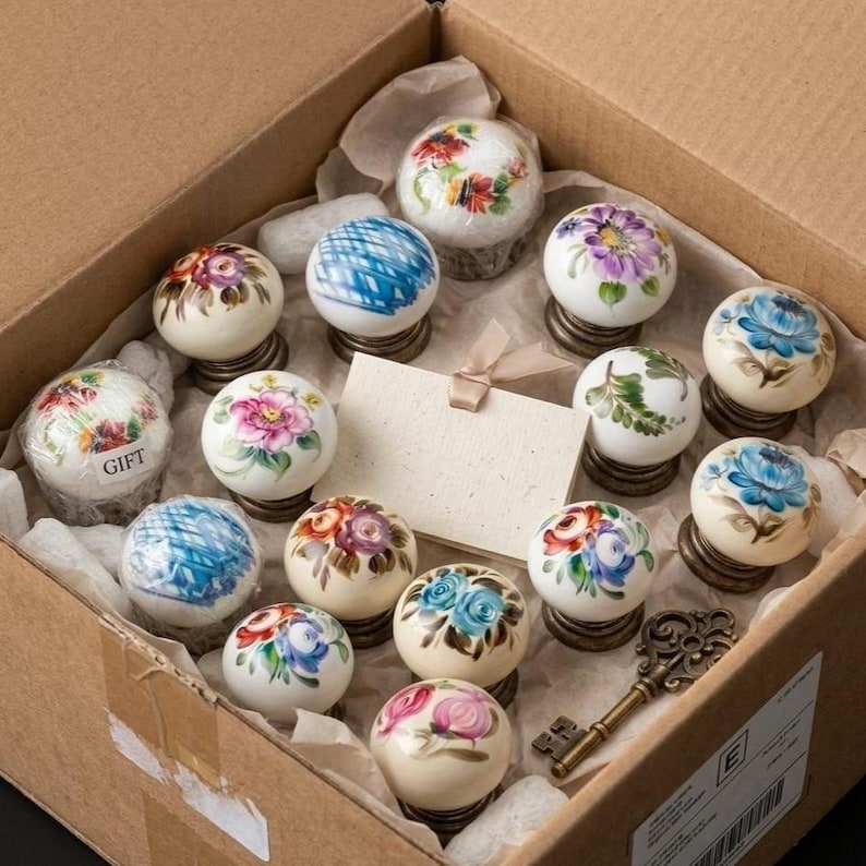 May include: A collection of round ceramic drawer knobs with hand-painted floral and geometric patterns in various colors, including blue, pink, and purple. The knobs feature metallic bases and are arranged in a cardboard box with a gift tag.
