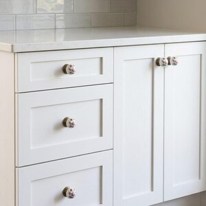 May include: White kitchen cabinetry featuring three stacked drawers and two side-by-side cabinet doors. Each drawer and door is fitted with a round white ceramic knob decorated with a small pink floral pattern and a metallic bronze-toned base.