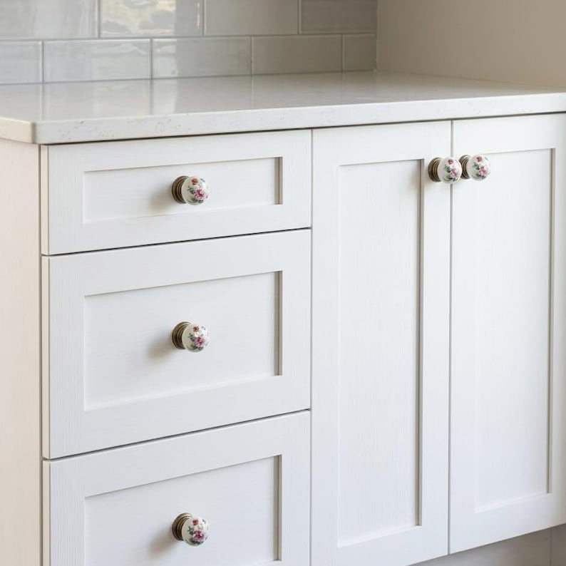 May include: White kitchen cabinetry featuring three stacked drawers and two side-by-side cabinet doors. Each drawer and door is fitted with a round white ceramic knob decorated with a small pink floral pattern and a metallic bronze-toned base.