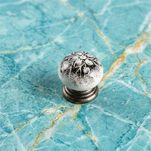 May include: A round ceramic cabinet knob featuring a white surface with a black floral pattern and a speckled gradient effect. The knob is attached to a circular, dark metallic base with a tiered, ridged design at the bottom of the fixture.