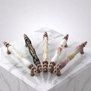 May include: Five cabinet pulls with antique bronze-toned metal ends and cylindrical ceramic centers. The designs include a white handle with gold accents, a black handle with gold filigree, a white handle with gold speckles, a white handle with a floral motif, and a textured handle with a purple gradient.