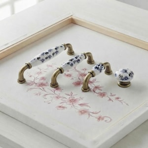 Peut inclure: Trois poignées de tiroir en métal et un bouton de placard rond avec des centres en céramique blanche à motifs floraux bleus et des ferrures en métal couleur laiton antique. Les éléments sont alignés sur une surface blanche à motif floral rose.
