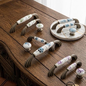 May include: A collection of ceramic drawer pulls and knobs featuring white surfaces with hand-painted floral patterns in green, blue, and purple. Each piece is accented with antique bronze-toned metal hardware, showcasing curved, ribbed ends for mounting on furniture.