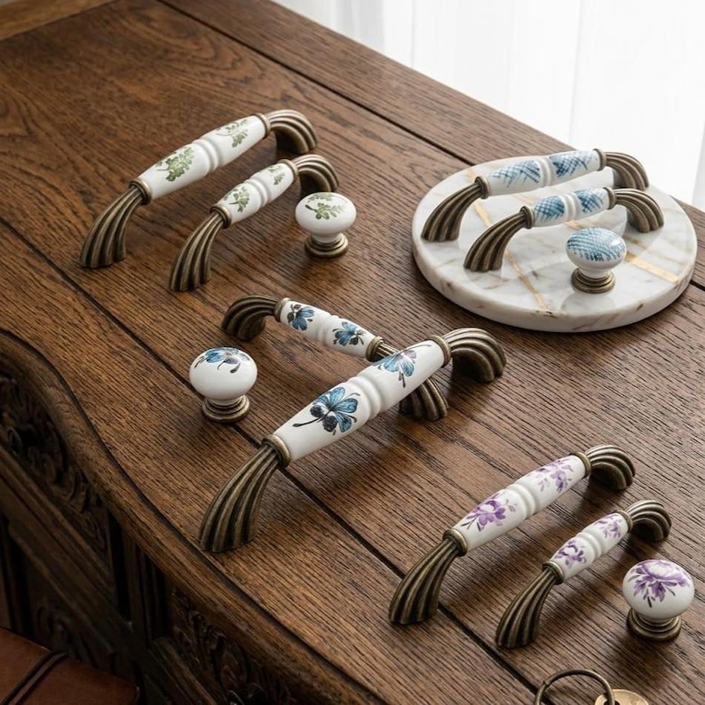 May include: A collection of ceramic drawer pulls and knobs featuring white surfaces with hand-painted floral patterns in green, blue, and purple. Each piece is accented with antique bronze-toned metal hardware, showcasing curved, ribbed ends for mounting on furniture.