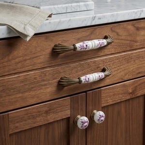 May include: Two horizontal drawer pulls and two round cabinet knobs installed on dark wood cabinetry. Each handle and knob features a white ceramic center with a purple floral pattern, flanked by fluted, antique brass-toned metal ends.