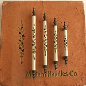 May include: Four cylindrical cabinet pulls of varying lengths with cream-colored bodies, dark metal finials, and hand-painted olive branch motifs. The pulls are mounted on a square terracotta tile featuring the text Modern Handles Co.
