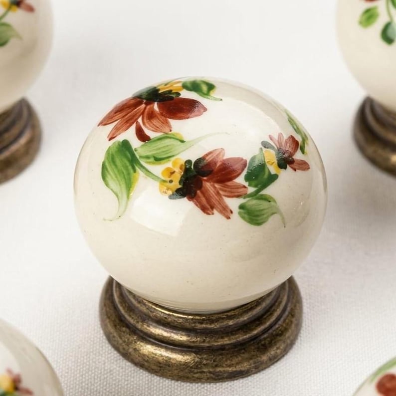 May include: A spherical ceramic cabinet knob with a cream-colored base featuring hand-painted floral motifs in shades of rust-red, yellow, and green. The knob is mounted on a circular, antique-finished brass base with a stepped, tiered design.