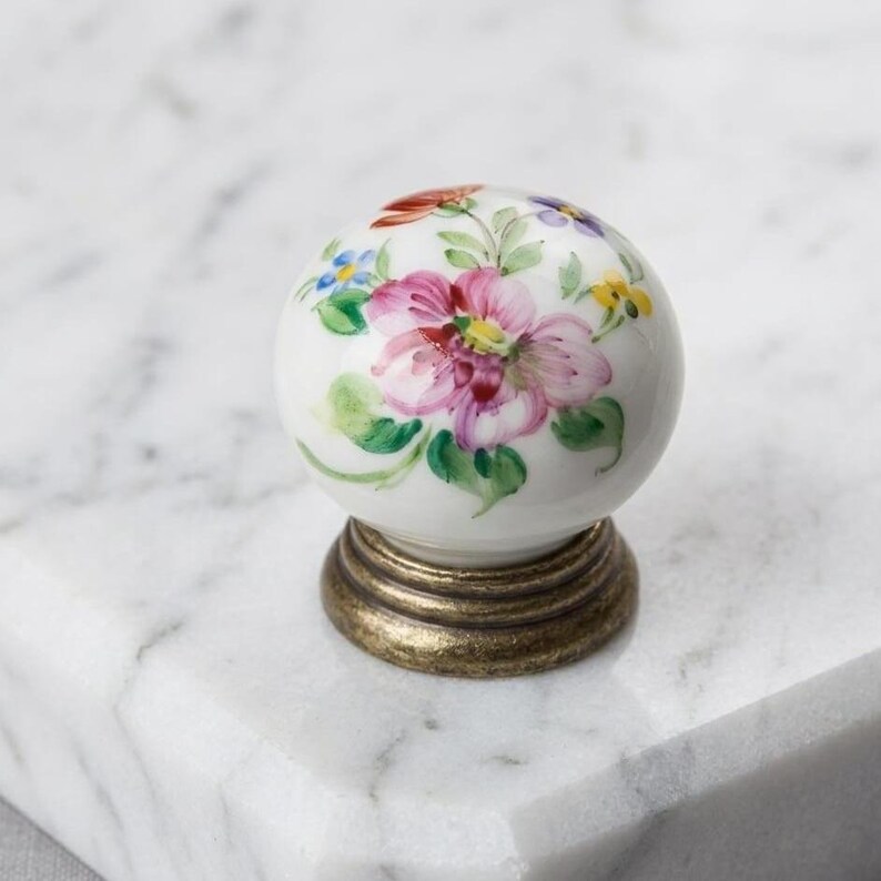 May include: A spherical white ceramic cabinet knob featuring a hand-painted floral design with pink, blue, yellow, and green accents. The knob is mounted on a circular, tiered base with a distressed, antique brass-toned metallic finish.