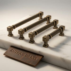 May include: Four antique brass cabinet pulls with a textured, knurled cylindrical grip and square mounting posts. The hardware is arranged in a row, and a brown leather tag with the embossed text ANTIQUE BRASS is placed in the foreground.