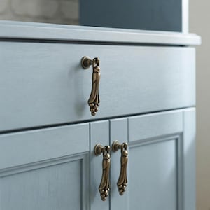 Könnte beinhalten: Drei antike Messing-Fallgriffe an einem hellblau lackierten Holzschrank. Jeder Griff hat ein vertikales, längliches Design mit einem abgerundeten oberen Befestigungspunkt und einer strukturierten, perlenartigen Basis, die einen Kontrast bildet.