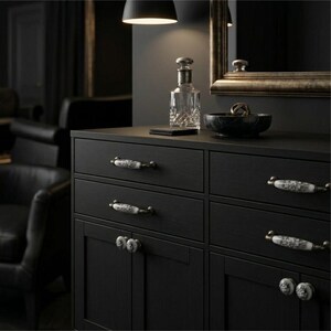 May include: A black wooden dresser featuring six drawers and two cabinet doors. Each drawer and door is equipped with ornate, white ceramic handles decorated with intricate floral patterns and metallic accents, providing a contrast.