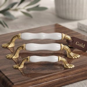 May include: Three cabinet pulls of varying lengths, each featuring a smooth white ceramic centre and ornate, brushed gold-tone metal ends with a fluted design. A small brown leather tag attached to the largest pull displays the word Gold.