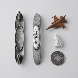May include: Two elongated cabinet pulls with a distressed silver finish, one featuring a central white oval inlay with a black floral motif. Also included are a small wooden toy airplane, a white ceramic bird figurine, and a round metal coin.
