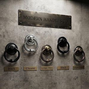 Peut inclure: Un panneau d'affichage étiqueté MODERN HANDLES CO présentant cinq poignées annulaires circulaires dans diverses finitions. La quincaillerie comprend deux noires, une chromée, une en laiton antique et une en cuivre antique, chacune avec une plaque.