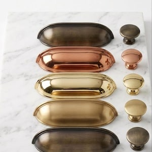 May include: A collection of five metal drawer pulls and five matching round knobs arranged in two vertical columns. The hardware features various metallic finishes including brushed bronze, polished copper, bright gold, satin brass, and dark antique brass.