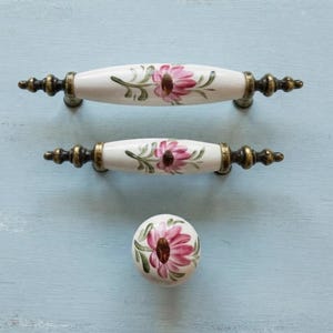 May include: Two white ceramic drawer pulls and one matching round knob, each featuring a hand-painted pink flower with green leaves and antique brass-toned metal hardware at the ends and base, arranged vertically on a light blue surface.