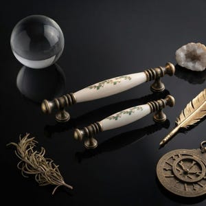 May include: Two cabinet pulls featuring a cream-colored ceramic center with a green leaf pattern and antique brass-toned metal ends. The pulls are arranged diagonally, accompanied by a glass sphere, a raw crystal, a gold feather, and a compass.