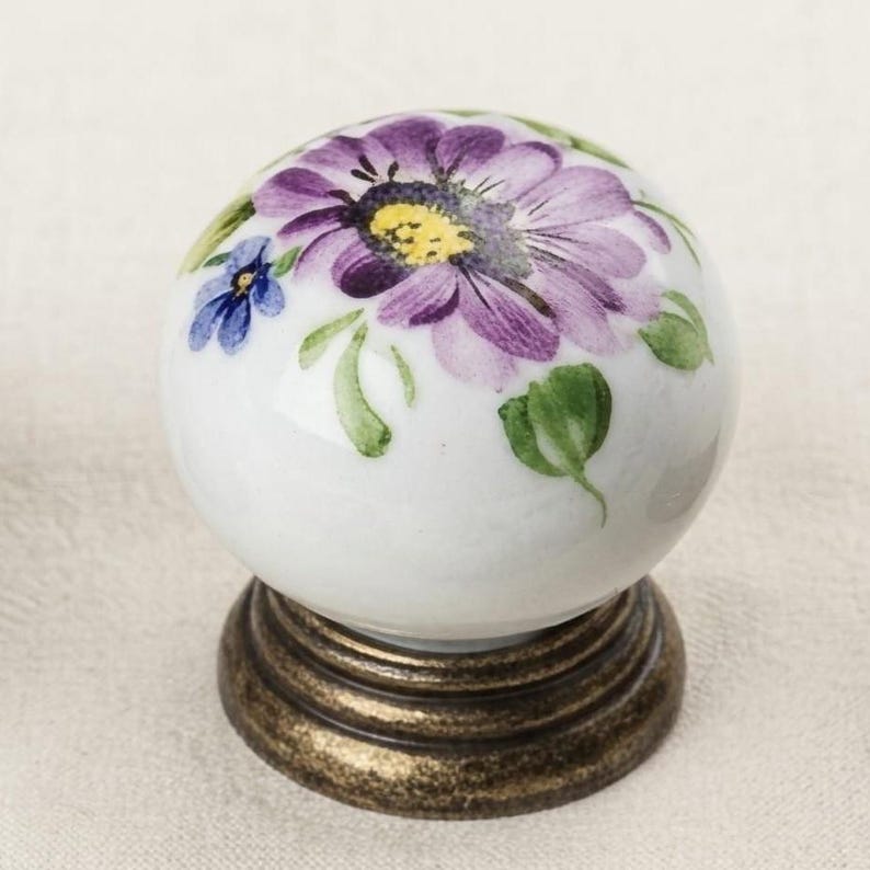 May include: A round white ceramic cabinet knob featuring a hand-painted purple flower with a yellow center, smaller blue flowers, and green leaves. The knob is mounted on a circular, tiered base with a distressed, antique bronze-colored finish.