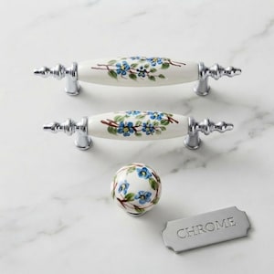 May include: Two elongated ceramic drawer pulls and one round ceramic knob, all featuring a white base with blue floral patterns and green leaves. The hardware includes polished chrome metal ends and a rectangular metal tag labeled CHROME.