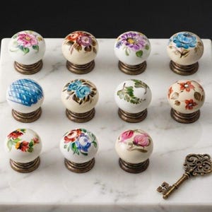 May include: A collection of eleven round ceramic cabinet knobs featuring various hand-painted floral and geometric patterns in multiple colors, each mounted on a metallic base, accompanied by a single ornate antique-style metal skeleton key.