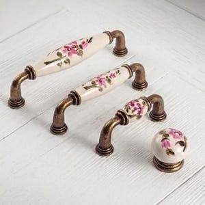 Peut inclure: Trois poignées de tiroir en céramique et un bouton rond en céramique, chacun présentant une base de couleur crème avec un motif floral rose peint et des feuilles vertes. La quincaillerie comprend des embouts et des bases en métal bronze antique.