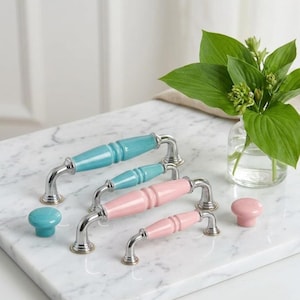 Può includere: Una collezione di ferramenta per mobili che include due maniglie per cassetti arcuate in turchese lucido e due in rosa pastello, ciascuna con estremità in metallo argentato. Due pomelli rotondi abbinati, uno turchese e uno rosa, sono posizionati ai lati.