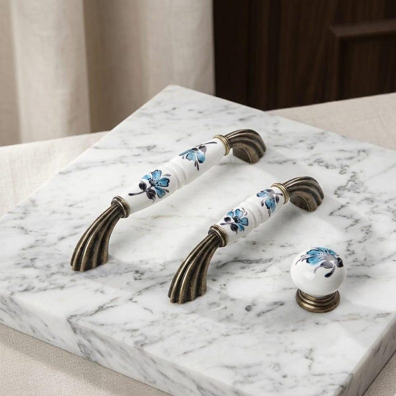 May include: Two arched cabinet handles and one round cabinet knob made of white ceramic with blue floral patterns and dark bronze-toned metal bases. The handles feature a ribbed design on the metal ends and a smooth, cylindrical ceramic center.