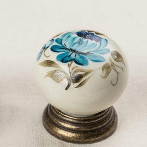 May include: A round, off-white ceramic cabinet knob featuring a hand-painted blue flower design with gold-toned leaves. The knob is mounted on a circular, dark metallic base with a tiered, ridged edge, providing a sturdy foundation for the piece.