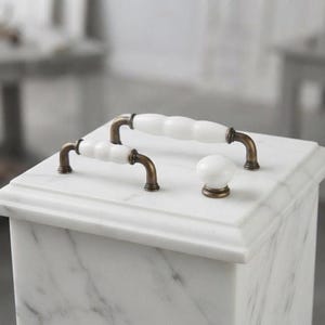 May include: Two white ceramic cabinet pulls with a scalloped, ribbed design and antique bronze metal ends, accompanied by a single matching white ceramic round cabinet knob with an antique bronze base, all resting on a white marble surface.
