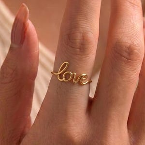May include: A gold-toned ring featuring the word love written in a continuous cursive script. The ring is constructed from a thin, smooth wire that forms the letters l, o, v, and e, creating a delicate and lightweight band that encircles the finger.