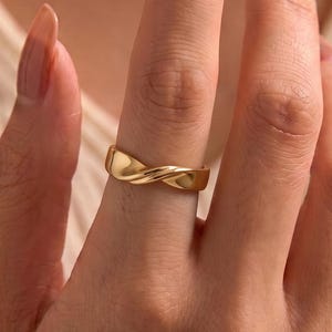 Op de afbeelding: Een gepolijste gouden ring met een glad, gedraaid bandontwerp. Het metaal heeft een reflecterend oppervlak dat het licht opvangt, waardoor subtiele highlights ontstaan langs de rondingen van de band. De ring wordt gedragen om de middelvinger.