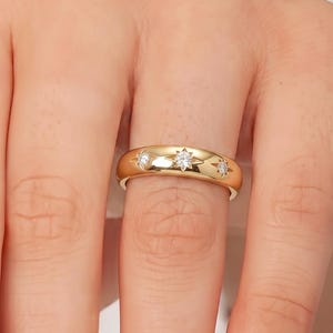 May include: A polished gold band ring featuring three small, clear, round-cut stones set in star-shaped engravings. The stones are arranged horizontally across the centre of the smooth, rounded metal band, creating a subtle, celestial-inspired design.