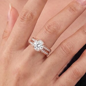 Puede incluir: Un conjunto de anillos de compromiso en oro rosa con una gran piedra central redonda de talla brillante sujeta por cuatro garras, flanqueada por dos bandas a juego. Las bandas están incrustadas con pequeñas piedras blancas en pavé.