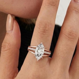 May include: A rose gold ring featuring a large marquise-cut centre stone and a split shank band. The band is adorned with small, clear pavé-set stones along the sides, creating a sparkling effect on the metal surface of the ring.