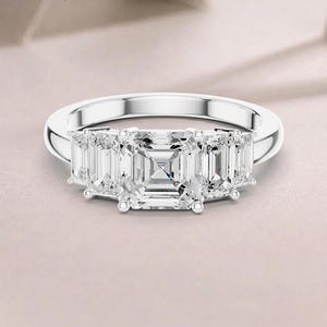 Puede incluir: Un anillo de tono plateado con una gran piedra central de corte cuadrado flanqueada por dos piedras más pequeñas de corte rectangular a cada lado, sujetas por garras metálicas sobre una banda lisa y pulida, creando un diseño de cinco piedras.