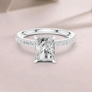 Op de afbeelding: Een verlovingsring met een radiant-geslepen diamant. De ring heeft een grote, rechthoekige diamant als middelpunt, gezet in een klauwzetting. De band is gemaakt van wit metaal en is bezet met kleinere diamanten.