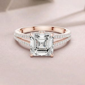 Puede incluir: Un anillo de oro rosa con una gran piedra central cuadrada y transparente. La banda se divide en dos brazos delicados, cada uno incrustado con una hilera de pequeñas piedras redondas y transparentes que se extienden desde el centro.