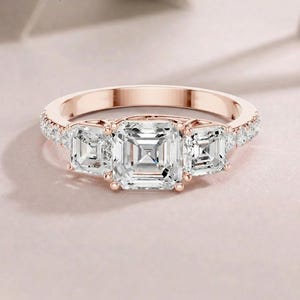 Puede incluir: Un anillo de oro rosa con una piedra central grande de corte cuadrado flanqueada por dos piedras más pequeñas de corte cuadrado. La banda está acentuada con una hilera de pequeñas piedras redondas engastadas en pavé en los laterales.