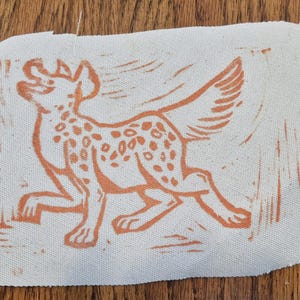 May include: A rectangular off-white fabric patch featuring a block-printed illustration of a spotted hyena in a rust-orange ink. The hyena is depicted in a dynamic, running pose with its head turned upward, showcasing a textured, hand-carved aesthetic.