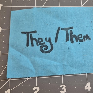 May include: A small, rectangular piece of light blue fabric with the words "They / Them" written in black, handwritten-style lettering. The fabric is placed on a gray cutting mat with grid lines.