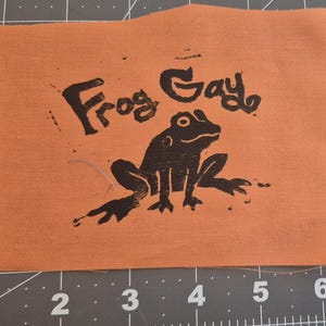 May include: Orange fabric square with the words "Frog Gay" in black, above a black frog illustration. The frog is sitting with its legs spread. The fabric is on a grey cutting mat.