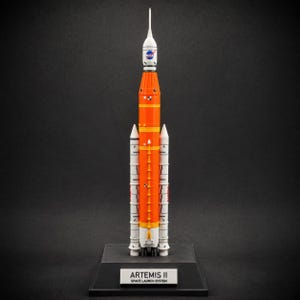 Pode incluir: Um modelo em escala do foguete NASA Artemis II Space Launch System. O foguete apresenta um estágio central laranja, propulsores laterais brancos e um estágio superior branco com o logotipo da NASA. Fica em uma base preta com uma placa lendo ARTEMIS II SPACE LAUNCH SYSTEM.