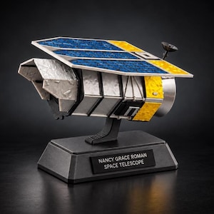 May include: A scale model of the Nancy Grace Roman Space Telescope featuring a large blue and yellow solar array, a silver metallic body, and a small black antenna. The model is mounted on a black rectangular base with a plaque reading NANCY GRACE ROMAN SPACE TELESCOPE.