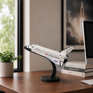 Puede incluir: Un modelo de transbordador espacial blanco montado sobre un soporte negro curvado. El transbordador presenta el texto Discovery, United States, una pequeña bandera estadounidense y el logotipo de la NASA. La cabina es negra y la aleta es blanca y negra.