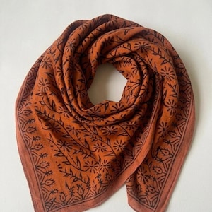 Pode incluir: Um lenço quadrado em tom de laranja queimado com um padrão floral estampado em bloco castanho escuro. O design inclui um motivo central de flores e vinhas estilizadas, emoldurado por uma borda distinta de formas botânicas repetidas.