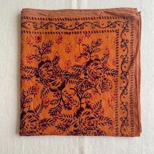 May include: A square fabric bandana in a burnt orange hue featuring a dark brown block-printed floral pattern. The design includes a central arrangement of roses and leaves surrounded by a decorative border of scrolling vines and geometric lines.